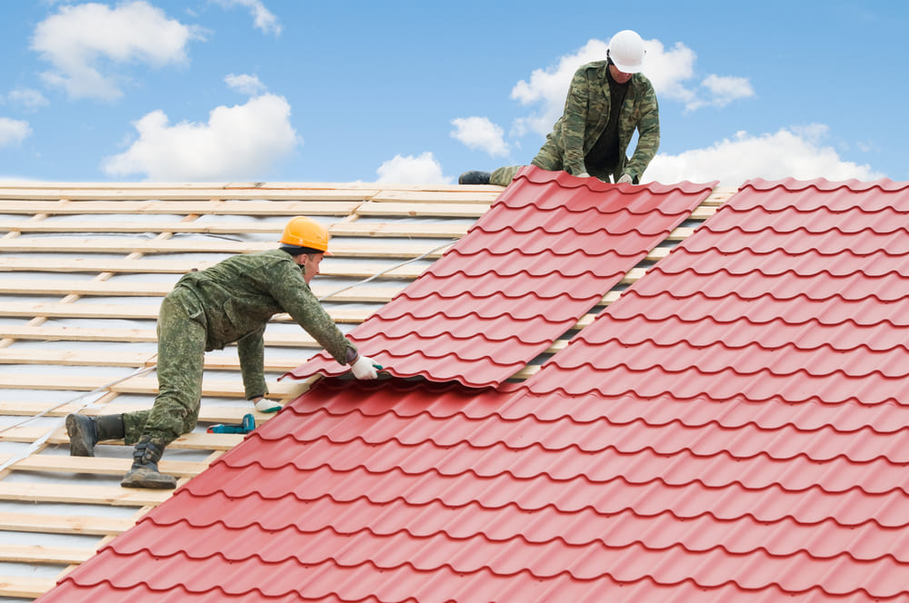 Two workers in hardhats install sheets of red metal roofing on top of a roof. 