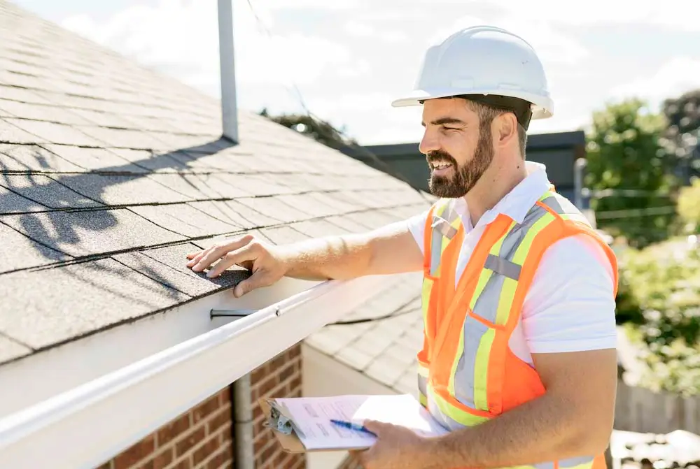 a roofer wearing a hard hat inspecting a roof while standing on a ladder